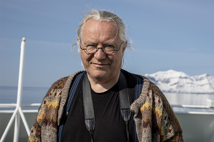 Mann står på dekket av en båt og ser i kamera, med fjell i bakgrunnen.